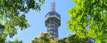 Petrin Lookout Tower viewed through a canopy of lush green trees on Petrin Hill in Prague, Czech Republic, under a clear blue sky