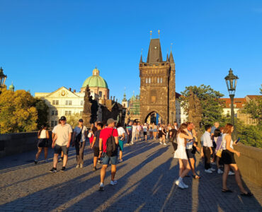 Crowds of tourists walking across the cobblestone Charles Bridge towards the Gothic Old Town Bridge Tower during golden hour in Prague.