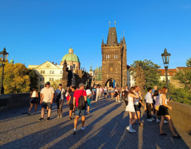 Crowds of tourists walking across the cobblestone Charles Bridge towards the Gothic Old Town Bridge Tower during golden hour in Prague.
