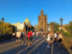 Crowds of tourists walking across the cobblestone Charles Bridge towards the Gothic Old Town Bridge Tower during golden hour in Prague.